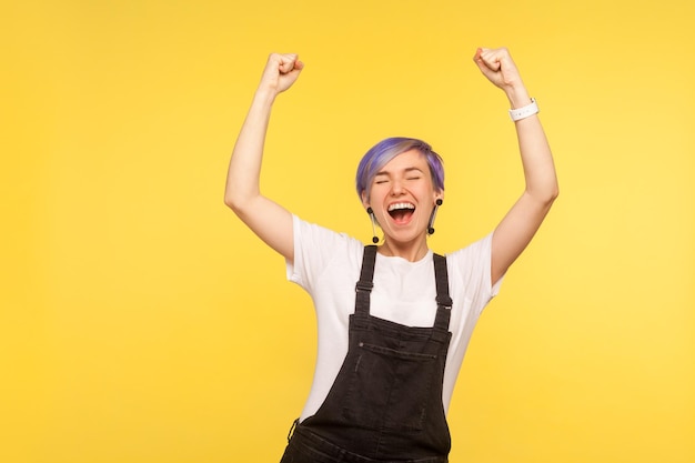 Portrait of ecstatic joyful hipster girl with violet short hair in denim overalls standing with her fists raised and shouting enthusiastically lively energetic winner celebrating victory studio shot