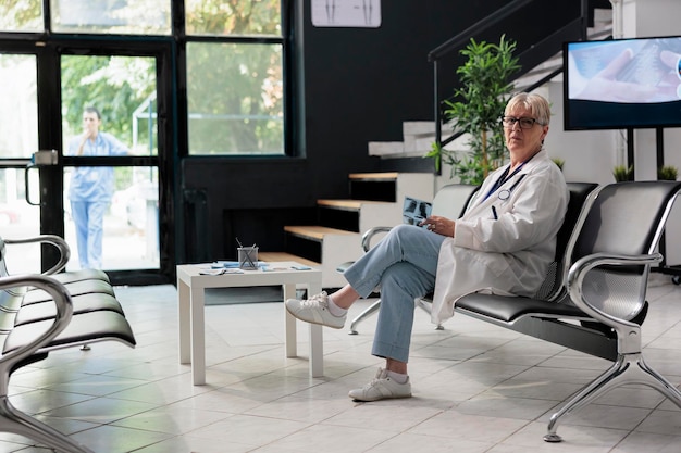 Portrait of elderly doctor sitting in hospital lobby waiting to attend medical examination with patients. Senior physician holding radiography exam scan. Medicine concept