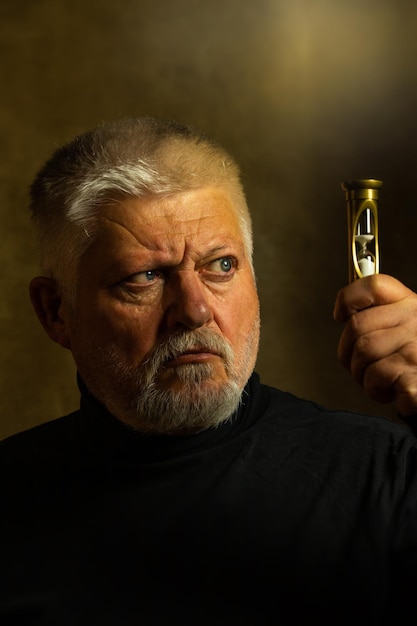 Photo portrait of an elderly man with a beard he looks skeptically at an hourglass