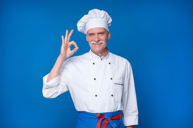 Portrait of elderly master chef isolated on blue wall, with okay gesture