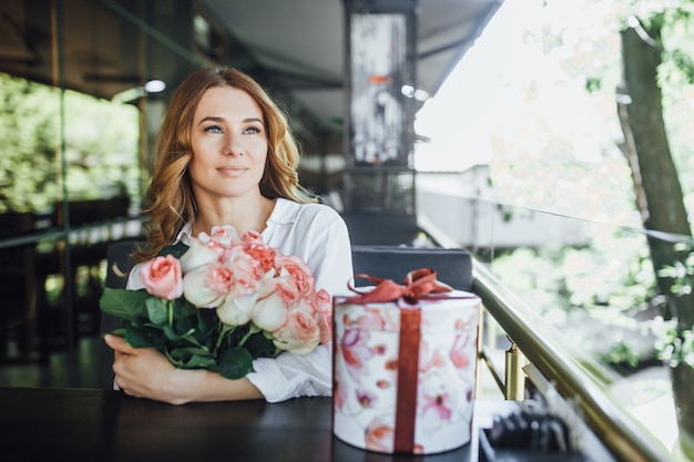 Portrait of an elderly woman holding a bouquet of roses and a gift on the summer terrace of a modern cafe.