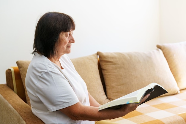 Portrait of an elderly woman reading book on couch at home caucasian pensioner spends her free time ...