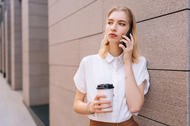 Portrait of elegant blonde female teenager in casual clothes holding takeaway coffee cup and talking on smartphone standing on city street in sunny summer day