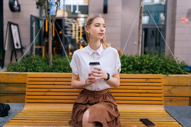 Portrait of elegant blonde young woman sitting on bench holding paper cup of takeaway coffee pensive looking away