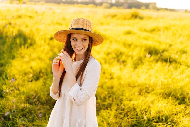 Portrait of elegant happy redhead woman wearing straw hat and white dress standing posing with closed eyes on beautiful meadow of green grass