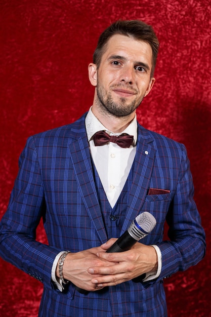 Portrait of elegant showman in blue suit and bow tie standing with microphone on red background