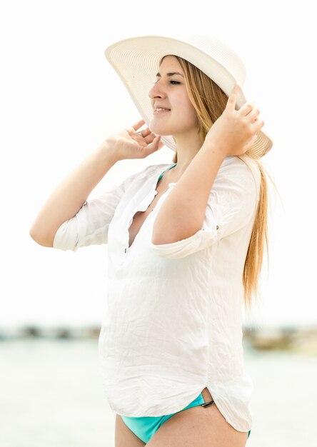 Portrait of elegant woman in hat standing on beach and looking at horizon