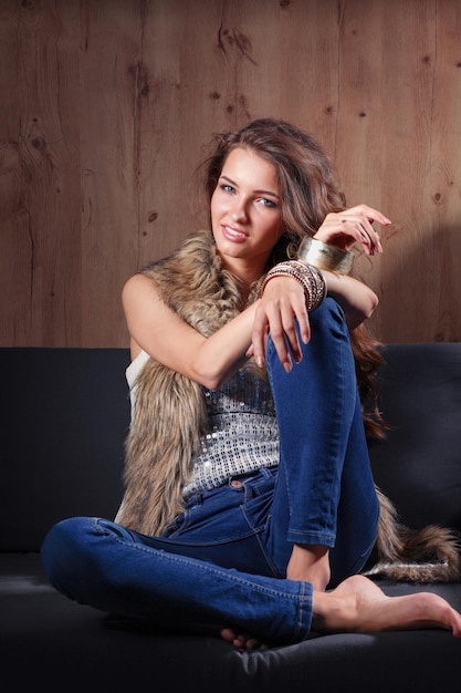 Portrait of elegant woman sitting on black sofa wearing a blue jeans and fur vest