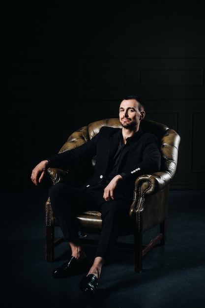 Photo portrait of an elegant young european man in a suit sitting on a chair in a dark interior.