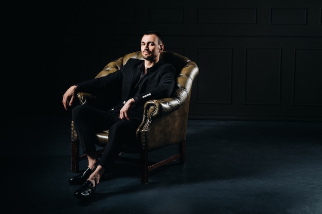 Portrait of an elegant young European man in a suit sitting on a chair in a dark interior.