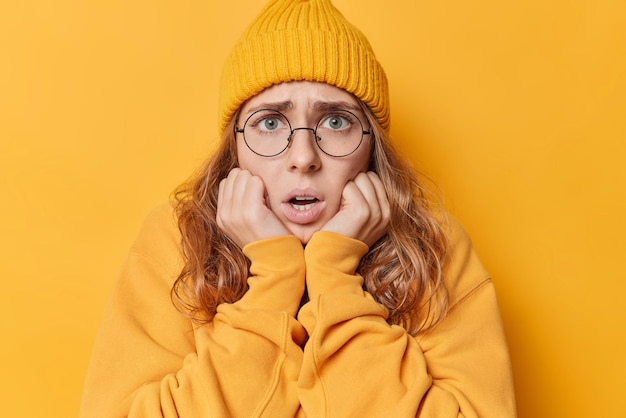 Portrait of embarrassed shocked young woman stares nervous keeps hands under chin jaw dropped being in panic dressed in casual clothes isolated over vivid yellow background. People and reactions