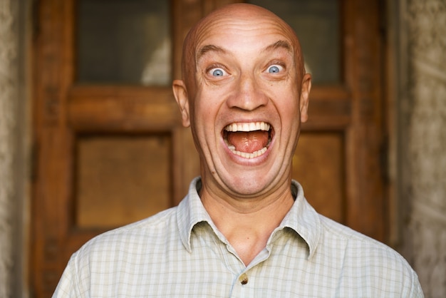 Portrait of an emotional bald guy with blue eyes dressed in light shirt shows different grimaces clo...