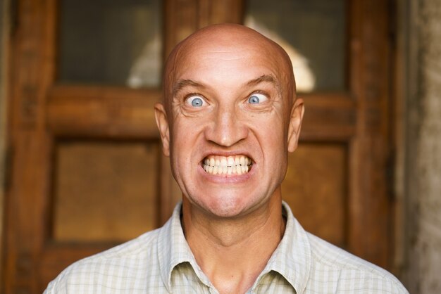 Portrait of an emotional bald guy with blue eyes dressed in light shirt shows different grimaces clo...