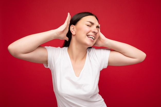 Portrait of emotional young attractive pretty brunette woman with sincere emotions wearing white t-shirt for mockup isolated over red background with empty space and covering ears with hands.