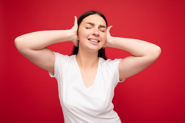 Portrait of emotional young attractive pretty brunette woman with sincere emotions wearing white t-shirt for mockup isolated over red background with empty space and covering ears.