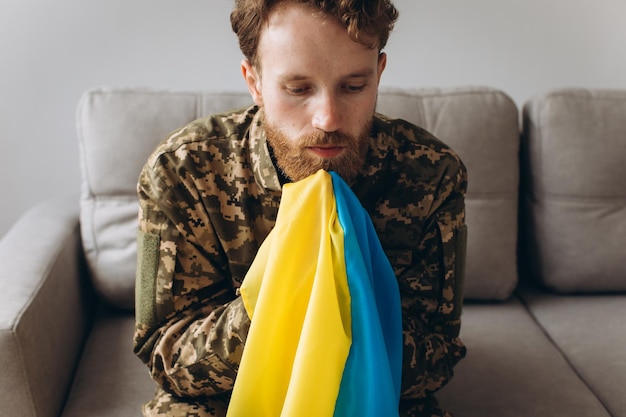 Portrait of an emotional young Ukrainian patriot soldier in military uniform sitting on the office on the couch holding a yellow and blue flag