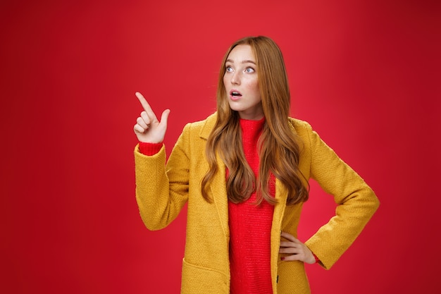 Portrait of empathic shocked and surprised stunned redhead woman in yellow coat open mouth amazed and speechless pointing and looking at upper left corner concerned and shook over red wall.