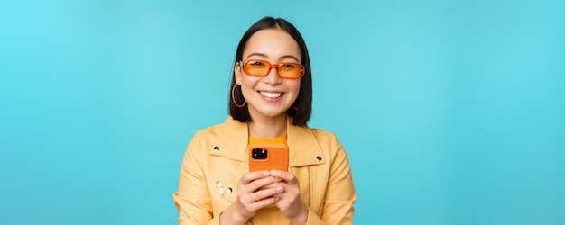 Portrait of enthusiastic asian woman in sunglasses using mobile phone smiling and laughing looking h