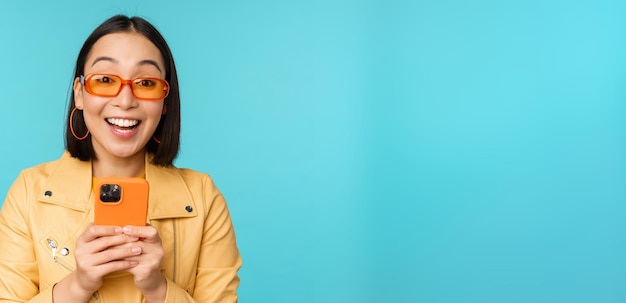 Portrait of enthusiastic asian woman in sunglasses using mobile phone smiling and laughing looking happy holding smartphone standing over blue background