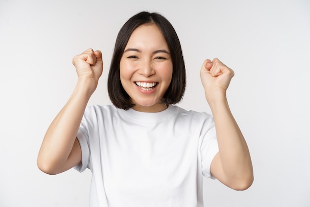 Portrait of enthusiastic asian woman winning celebrating and triumphing raising hands up achieve goal or success standing over white background