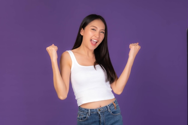 Portrait of excited Beautiful Asian brunette woman cute girl in white tank top raising hand in the air laughing cheerfully gesturing for winner smiling look camera isolated purple background