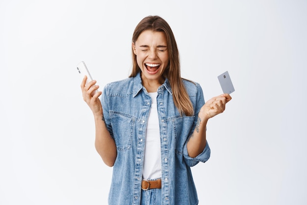 Portrait of excited blond girl screaming of happiness, winning money, holding plastic credit card and smartphone, standing on white