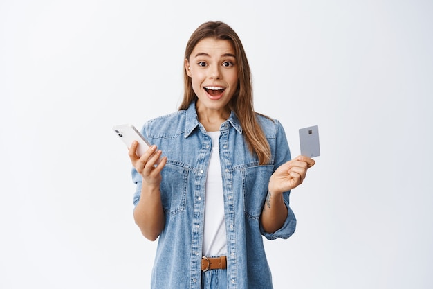 Portrait of excited blond girl shopping online on smartphone, holding credit card and smiling amazed, standing over white wall