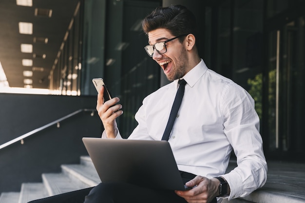 Portrait of excited businessman dressed in formal suit sitting outside glass building with laptop, and holding smartphone