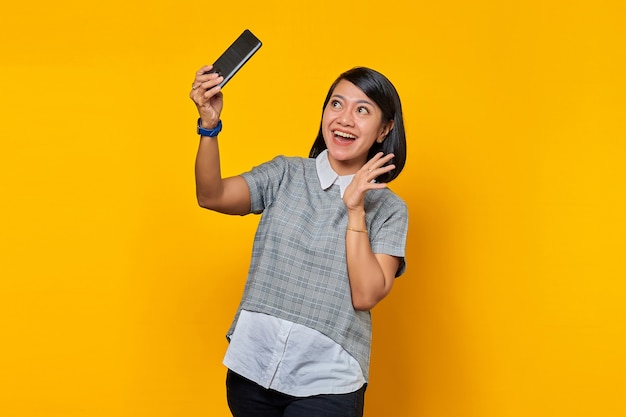 Portrait of excited cheerful young Asian woman using mobile phone take selfie over yellow background