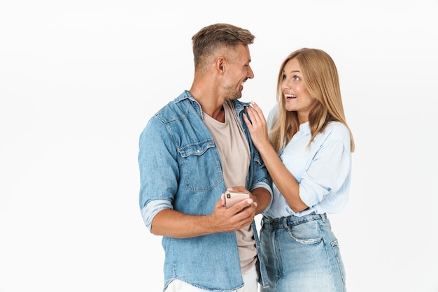 Portrait of excited couple man and woman in denim clothing smiling and using smartphone together isolated on white