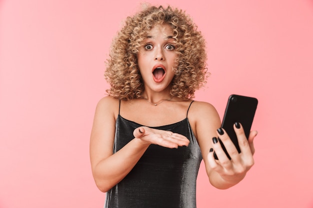 Portrait of excited curly woman 20s smiling and taking selfie photo on smartphone