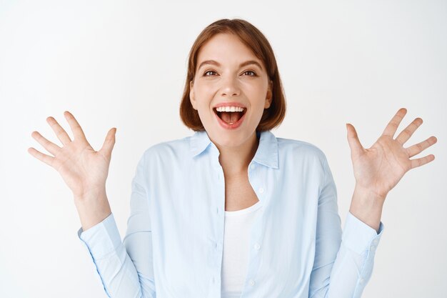 Portrait of excited and enthusiastic woman, spread hands sideways and scream of joy, winning, feeling happy and upbeat, celebrating on white wall