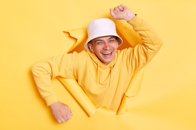 Portrait of excited funny man wearing casual hoodie and panama looking through torn hole in yellow paper looking at camera having arms raised arms dancing