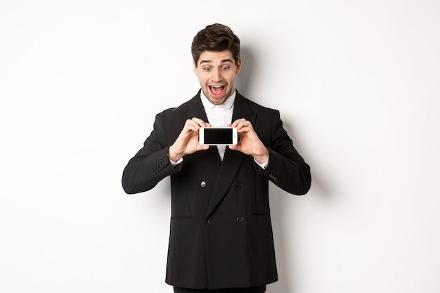 Portrait of excited handsome man showing smartphone screen, looking amazed at display with advertisement, standing over white background in black suit