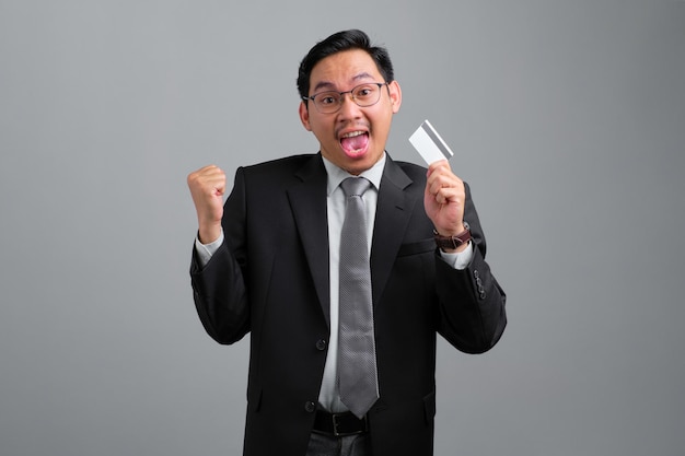 Portrait of excited handsome young businessman in formal suit holding credit card and doing winner gesture isolated on grey background