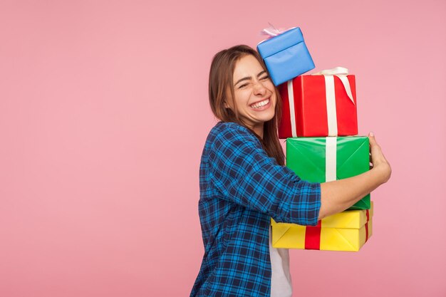 Portrait of excited happy girl in checkered shirt embracing lot of christmas gifts and smiling joyfully, celebrating birthday, admiring many presents. indoor studio shot isolated on pink background