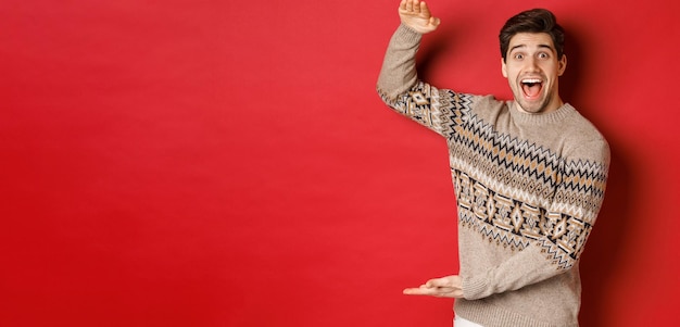 Portrait of excited and happy handsome man in christmas sweater showing something big holding large gift for holidays standing over red background