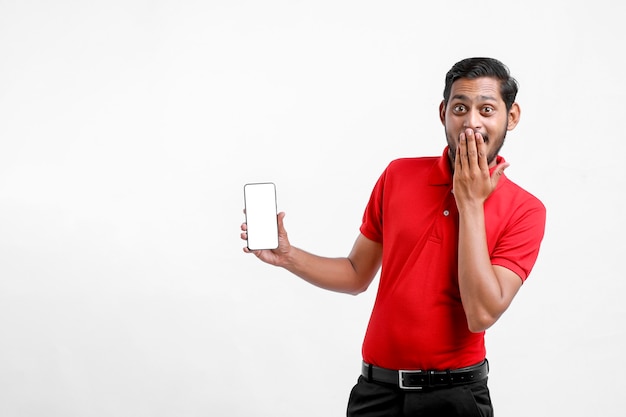 Portrait of a excited happy young delivery man in red t shirt and showing smartphone over white background.