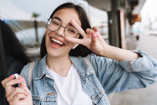 Portrait of a excited happy young pretty woman student wearing eyeglasses walking outdoors resting listening music with earphones showing peace gesture