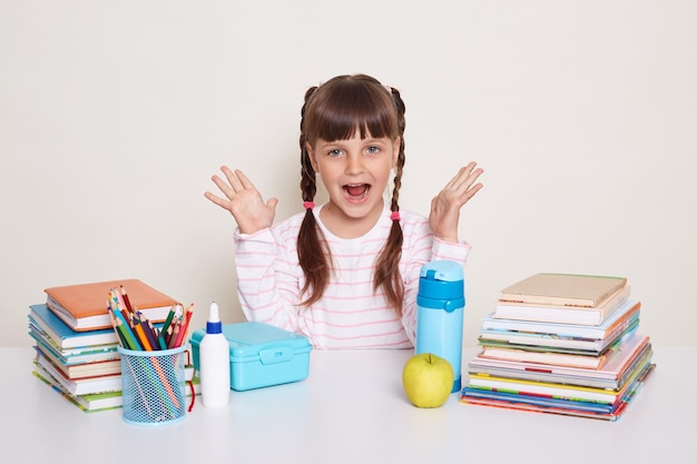 Portrait of excited positive little schoolgirl with pigtails wearing striped shirt sitting at the desk surrounded with books and other school supplies raised arms and screaming happily