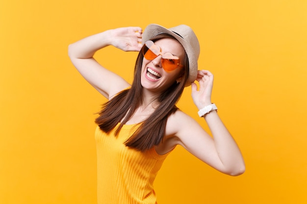 Portrait of excited smiling young woman in straw summer hat, orange glasses put hands on head, look aside isolated on yellow background. People sincere emotions, lifestyle concept. Advertising area.