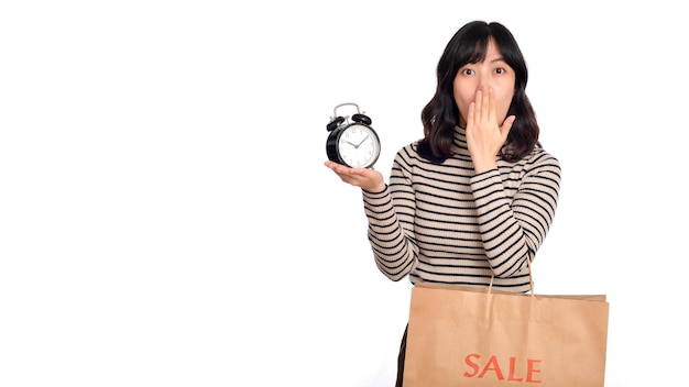 Portrait of excited young Asian woman with sweater shirt holding alarm clock and shopping paper bag isolated on white background last deals shocked price shopping concept