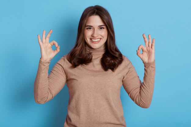 Portrait of excited young attractive girl looking directly with charming smile, showing ok signs with both hands, wearing beige turtleneck, posing isolated over blue wall.