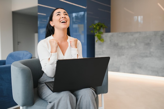 Portrait of excited young caucasian woman celebrating success while sitting with laptop at home or modern office Happy female freelancer working in coworking space