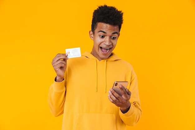 Portrait of an excited young teenager boy standing isolated over yellow wall, showing credit card while using mobile phone