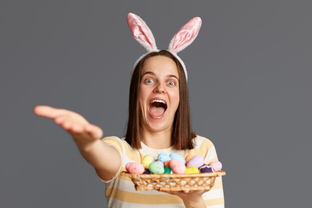 Photo portrait of extremely happy overjoyed woman wearing rabbit ears holding colorful easter eggs isolated on gray background outstretching hand inviting you to celebrate holiday together