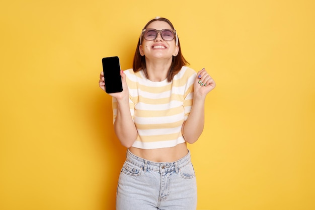 Portrait of extremely happy woman wearing sunglasses striped Tshirt and hair band posing isolated over yellow background showing mobile phone with blank black screen and clenched fist