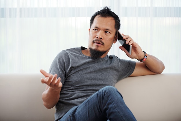 Portrait of eyoung man in tshirt and jeans resting on sofa and chatting on phone