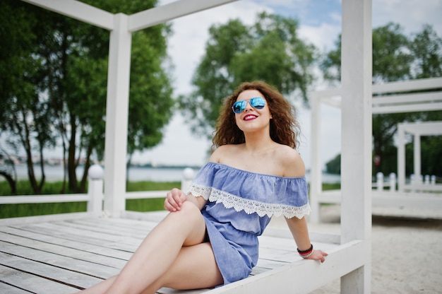 Portrait of a fabulous young woman wearing chic outfit posing on a white wooden terrace.