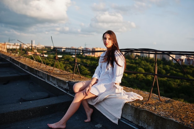 Portrait of a fabulous young woman in white male shirt sitting on the edge of the building roof with a fantastic view of a city in a background